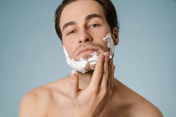 Attractive young smiling nude man with shaving foam on his face isolated over blue background - Foto, immagini