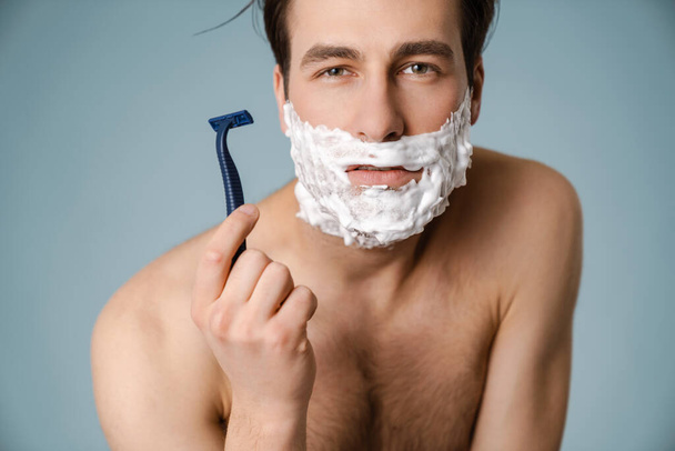 Shirtless white man looking at camera while shaving with foam isolated over blue wall - Foto, Imagem
