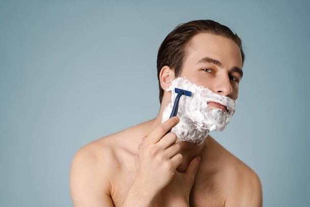 Attractive young smiling nude man with shaving foam on his face isolated over blue background, holding razor - Fotó, kép