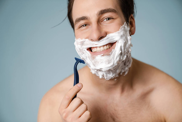 Shirtless white man smiling while shaving with foam isolated over blue wall - Fotó, kép