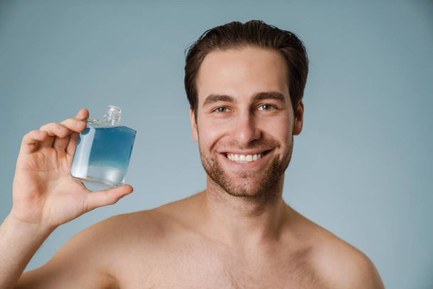 Shirtless white man smiling while showing cologne isolated over blue wall - Foto, imagen