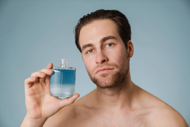 Shirtless white man looking at camera while showing cologne isolated over blue wall - Fotografie, Obrázek