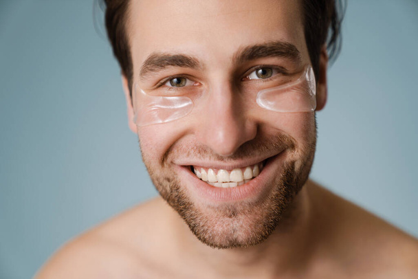 Shirtless white man with under eye patches smiling at camera isolated over blue wall - Fotografie, Obrázek