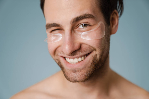 Shirtless white man with under eye patches winking at camera isolated over blue wall - Fotoğraf, Görsel