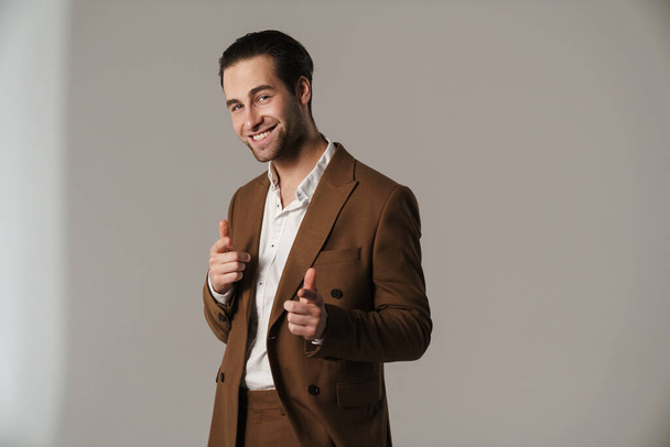 Unshaven brunette man in jacket smiling and pointing finger at camera isolated over grey background - Foto, afbeelding