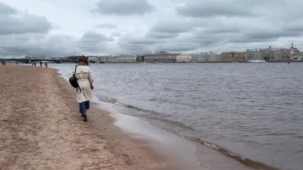 Молодая женщина гуляет в облачную погоду вдоль набережной Невы в Санкт-Петербурге с видом на Дворцовую площадь. - Кадры, видео