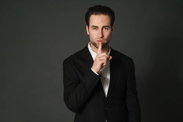 Unshaven brunette man in jacket looking and showing silence gesture isolated over black background - Φωτογραφία, εικόνα