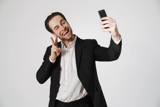 Brunette man showing peace sign and taking selfie on cellphone isolated over white background - Fotoğraf, Görsel