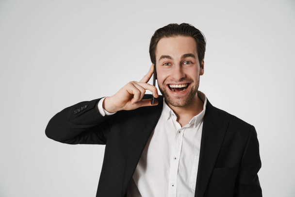 Unshaven brunette man in jacket smiling while talking on cellphone isolated over white background - Zdjęcie, obraz