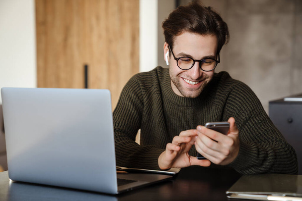 Smiling brunette man using phone and laptop computer while sitting at tbe table at home - Foto, Bild