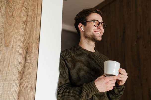 Smiling mid aged brunette man in earphones standing holding cup indoors - Фото, изображение