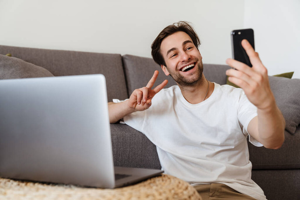 Smiling brunette man sitting on the floor with laptop computer, using mobile phone - Fotó, kép