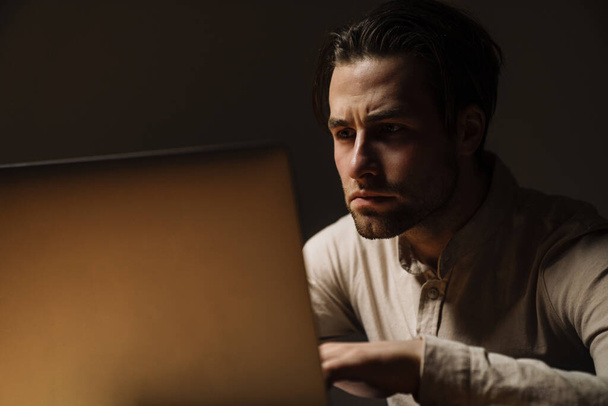 Serious pensive brunette businessman working on laptop computer in dark office - Zdjęcie, obraz