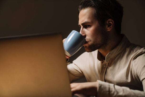 Serious pensive brunette businessman working on laptop computer in dark office - Foto, afbeelding