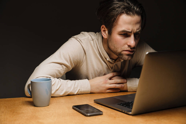 Serious pensive brunette businessman working on laptop computer in dark office - Фото, изображение
