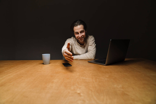 Smiling brunette businessman working on laptop computer in dark office, reaching his hand for a handshake - Foto, imagen