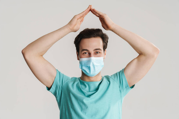 Caucasian pleased guy in protective mask showing home gesture isolated over white wall - Foto, Imagen