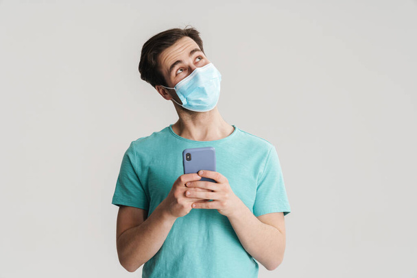 Joyful handsome guy in protective mask using mobile phone isolated over white wall - Foto, Imagen