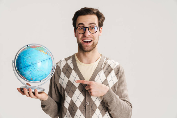 Geeky Hipster zeigt auf Globus und lächelt in Kamera auf grauem Hintergrund - Foto, Bild