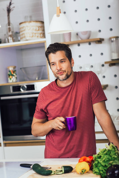 Handsome young man in casual wear drink coffee while standing in the kitchen at home - Photo, image