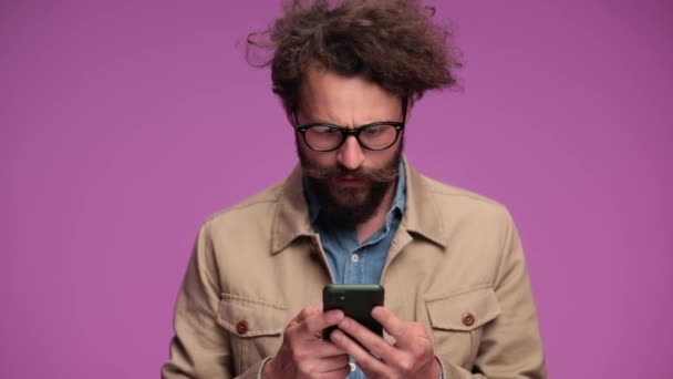 σοκαρισμένος σγουρά μαλλιά άνθρωπος κρατώντας κινητό, ανάγνωση ειδήσεων, να εκπλαγείτε και να πάρει απογοητευμένος καλύπτει το στόμα με το χέρι και γνέφοντας σε ροζ φόντο στο στούντιο - Πλάνα, βίντεο