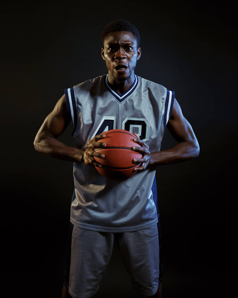 Agressieve basketbalspeler poseert met bal in de studio, zwarte achtergrond. Professionele mannelijke baller in sportkleding spelen sport spel, lange sporter - Foto, afbeelding