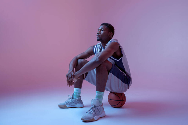 Basketball player sitting on ball in studio, neon background. Professional male baller in sportswear playing sport game, tall sportsman - Valokuva, kuva