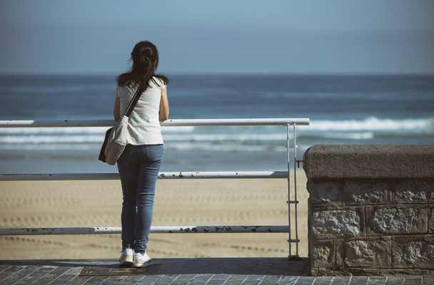 Güneşli bir günde, deniz kenarındaki metal çitlerin üzerinde duran esmer bir kadının arka görüntüsü. - Fotoğraf, Görsel