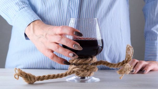 θηλυκό χέρι κρατώντας ποτήρι κρασί είναι δεμένο με ένα σχοινί γιούτα. Η έννοια της εξάρτησης από το αλκοόλ. - Φωτογραφία, εικόνα