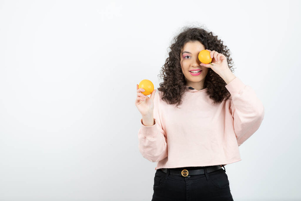Young curly woman in pink sweater holding oranges in front of eye. High quality photo - Fotografie, Obrázek