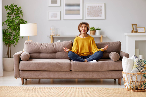 Peaceful african american teenage girl sitting   on couch and meditating at home. Young mixed race female student relaxing in lotus pose and relief stress - Fotoğraf, Görsel