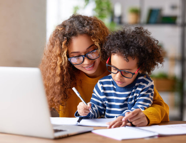 Дитина-афроамериканець пише щось у записнику з мамою, працюючи віддалено від дому на ноутбуці, допомагаючи маленькому синові домашнє завдання, сидячи вдома. Дитяча концепція - Фото, зображення