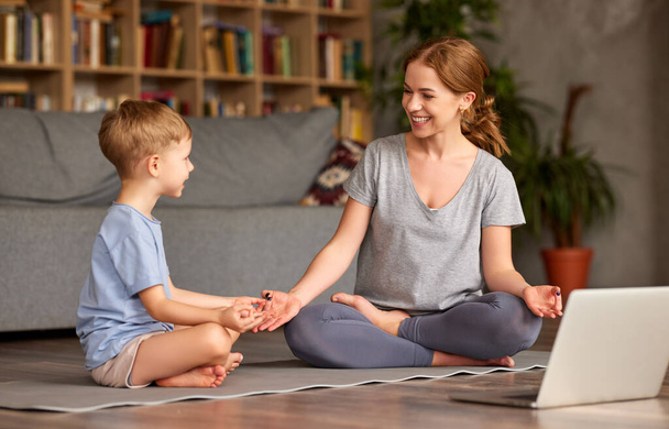 Gündelik giyinmiş genç güzel bir anne, küçük oğluna nilüferde birlikte otururken nasıl meditasyon yapacağını öğretirken evde oturma odasında mutlu bir aile, dizüstü bilgisayarla yoga egzersizleri yapıyor. - Fotoğraf, Görsel