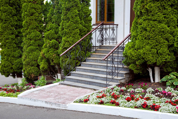 a granite staircase with a wrought iron railings, entrance steps, exit to the backyard with a flowerbed with flowers and evergreen thuja trees, nobody. - Photo, image