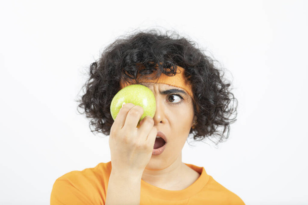 Sportowa kręcona kobieta w pomarańczowym stroju trzymająca zielone jabłko przed oczami. Wysokiej jakości zdjęcie - Zdjęcie, obraz