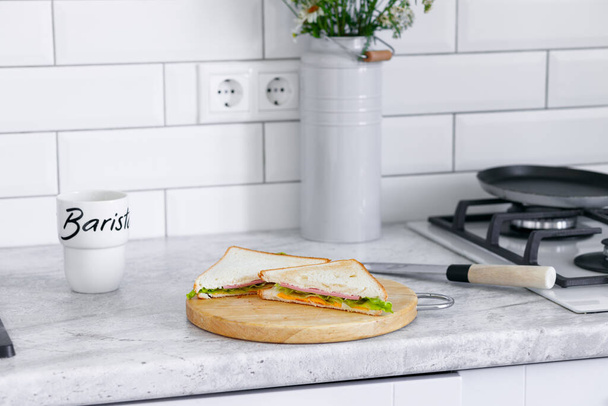Το κομμένο σάντουιτς βρίσκεται σε μια ξύλινη σανίδα στο τραπέζι της κουζίνας. Λευκό φόντο, πρωινό - Φωτογραφία, εικόνα