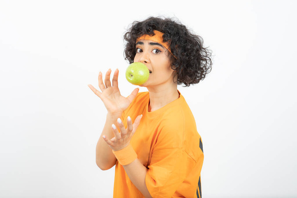 Deportiva mujer rizada en traje naranja sosteniendo manzana verde en su boca. Foto de alta calidad - Foto, imagen