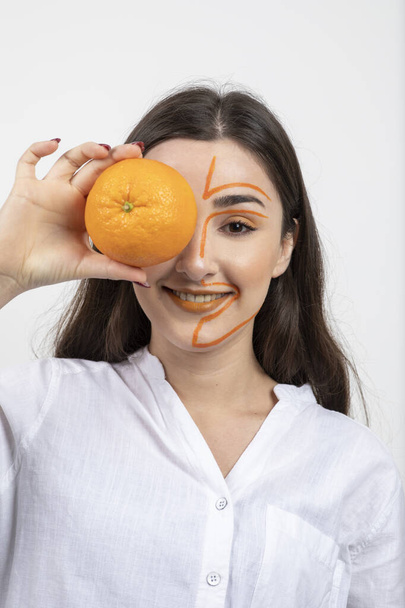 Imagen de mujer joven con la cara pintada cubriendo el ojo con fruta entera de color naranja. Foto de alta calidad - Foto, imagen