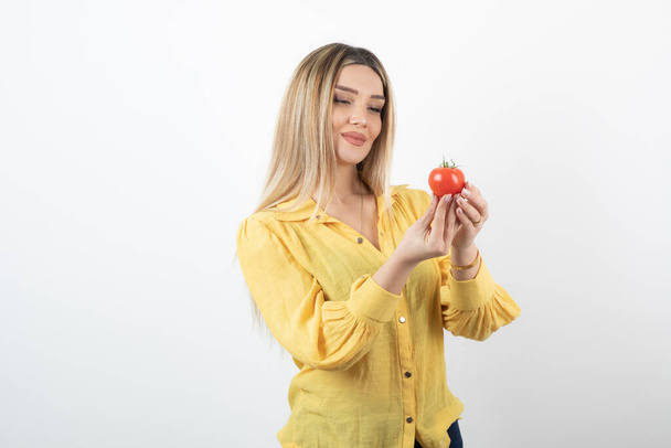 Young beautiful woman in yellow shirt looking at red tomato on white background. High quality photo - Фото, зображення