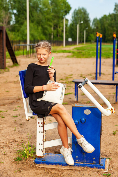 A blonde girl in a school black dress sits on a street simulator. White bag, white sneakers - Photo, Image
