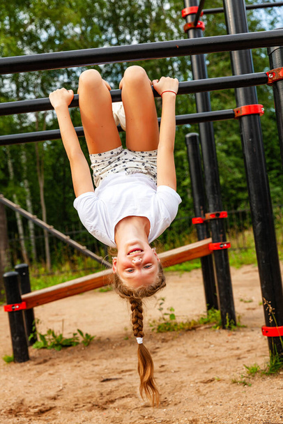 Una ragazza in uniforme sportiva pende a testa in giù sul tornello. Campo sportivo, ricarica, riscaldamento, giorno, aria aperta - Foto, immagini