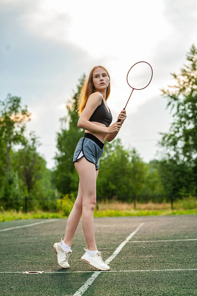 Νεαρή όμορφη κοπέλα με αθλητικό μπλουζάκι και σορτς στο γήπεδο. Μέρα, γυμναστήριο, υπαίθριος χώρος, μπάντμιντον, ρακέτα - Φωτογραφία, εικόνα