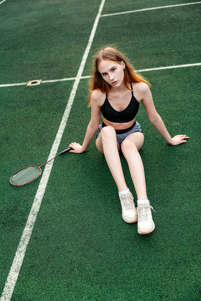 Fiatal gyönyörű lány sportfelsőben és rövidnadrágban ül a sportpályán. Nappali, fitness, szabadtéri, tollaslabda, ütő - Fotó, kép