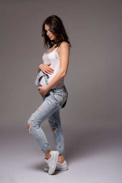 beautiful pregnant brunette girl posing in studio on gray background. Embracing the belly. in a white T-shirt and denim overalls. side view. vertical photography - Foto, Imagen