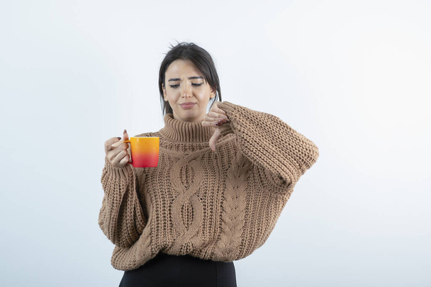 Image d'une belle femme en tricot tenant une tasse de thé orange et donnant les pouces vers le bas. Photo de haute qualité - Photo, image