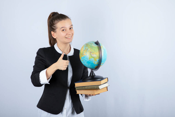 Kitapları ve küresi olan genç ve güzel bir öğretmenin beyaz arka planda baş parmağını kaldırdığı fotoğraf. Yüksek kalite fotoğraf - Fotoğraf, Görsel