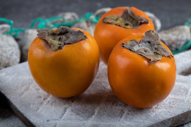 Dried persimmons with sugar powder around fresh persimmons on black surface. High quality photo - Фото, изображение