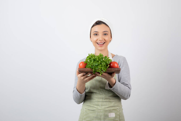 Zdjęcie uśmiechniętej modelki trzymającej drewnianą deskę ze świeżymi warzywami. Wysokiej jakości zdjęcie - Zdjęcie, obraz