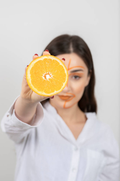 Obraz młodej kobiety z pomalowaną twarzą trzymającej połówkę pomarańczy. Wysokiej jakości zdjęcie - Zdjęcie, obraz