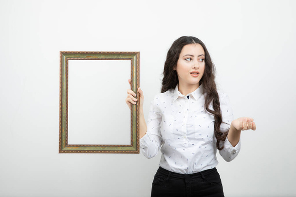Mujer joven de pie y sosteniendo el marco de la imagen contra la pared blanca. Foto de alta calidad - Foto, Imagen